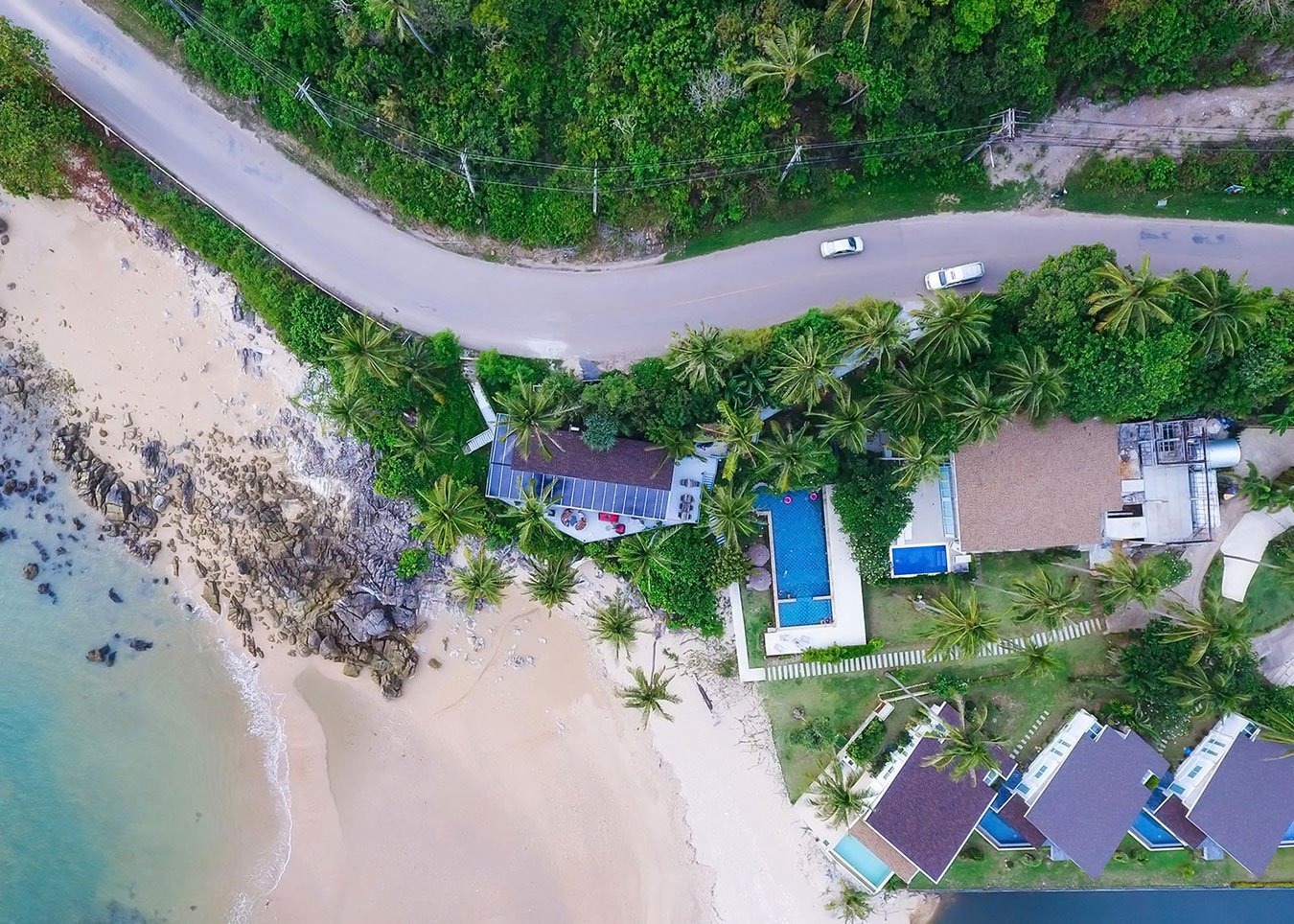 Vue aérienne de villas avec piscine en bord de plage bordée de palmiers et végétation tropicale luxuriante
